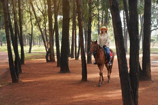 Young woman in shirt and straw hat, rides brown horse in the morning park, blurred trees in background