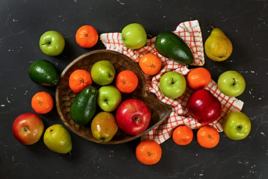 Apples, pears, oranges and avocado in wooden carved bowl on black marble like board, view from above