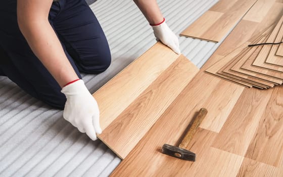 Installing laminated floor, detail on man hands in white gloves, holding wooden tile, over white foam base layer, small hammer near