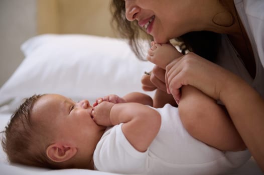 Close-up of a dark-haired multi ethnic young woman, a happy loving caring affectionate mother smiling to her lovely baby boy, kissing his tiny toes and feet. Childcare and maternity leave lifestyle