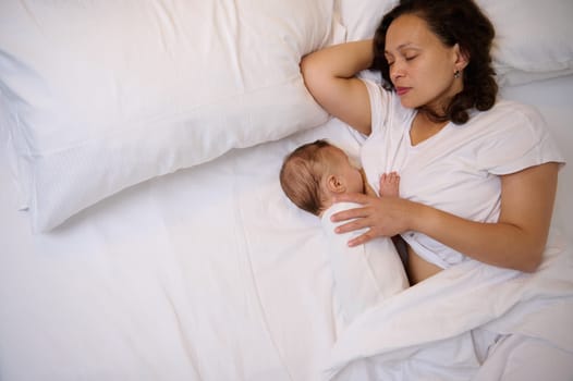 The concept of co-sleeping and breastfeeding. View from above of a sleeping mother breastfeeding her newborn baby, sleeping together on comfortable white bed. Maternity leave. Copy advertising space