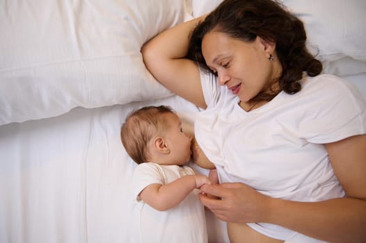 Overhead view of happy delightful young mom breastfeeding her cute newborn baby, lying together on the bed. Breastfeeding as a method of preventing breast cancer and the best nutrition for children