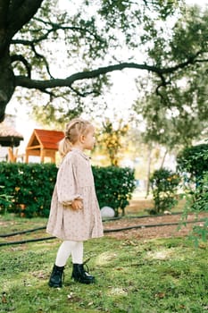 Little girl walks on a green lawn near a playground, looking to the side. High quality photo