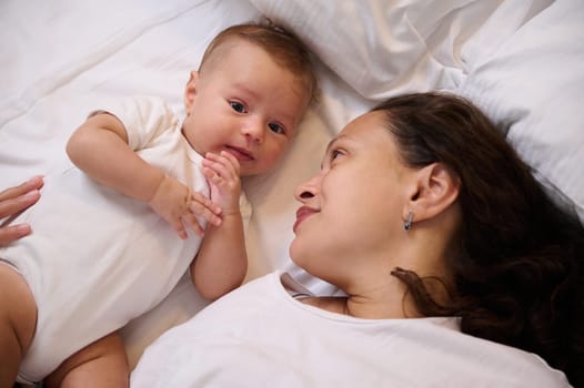 Happy young mother and child baby boy lying together on the bed, smiling, enjoying happy moments together. Family relationships. Babyhood. Child and baby care. Maternity leave and motherhood. Ad space