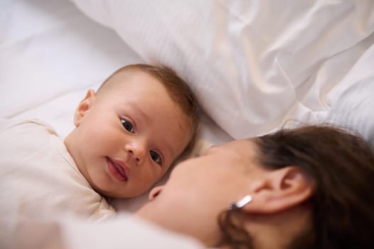 Close-up portrait of a cute baby boy, adorable Caucasian child 4 months old looking at camera, lying on the soft white bed sheets close to his loving affectionate mother. People. Two generation family