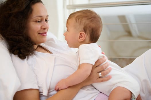 Newborn baby in tender embrace of mother. Happy delightful young smiling woman, affectionate loving mother caring of her newborn baby at home, lying together on the bed at cozy and light home interior