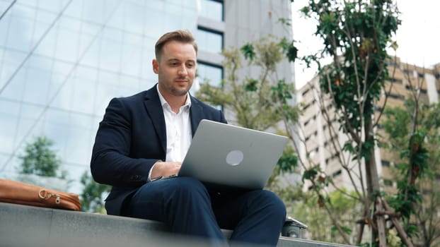 Skilled male leader working on his phone sending report while using laptop analyze investment data at green city. Happy investor looking marketing plan while sitting at park or eco urban city. Urbane.