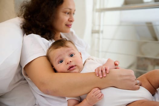 Authentic portrait of lovely baby boy 4 months old in the arms of her loving delightful mother dreamily looking away, enjoying happy time with her newborn child at home. Maternity leave. Infancy time