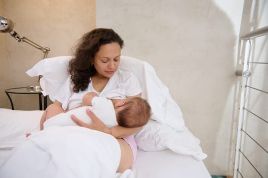 Young mother sitting on the bed and breastfeeding her baby. Maternity leave and infancy concept. People. Lifestyle. Mother's milk is the best nutrition for first 6 months of kids life