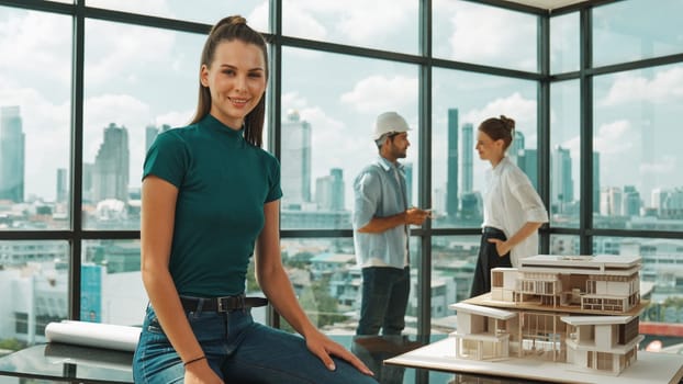 Young beautiful interior designer looking at camera while engineer team talking about house design. Skilled businesswoman smiling at camera with confident while sitting near house model. Tracery