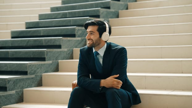 Project manager listening and enjoy music from headphone while sitting at stairs. Professional businessman wearing suit outfit while dancing and moving to music. Happy man listen funny song. Exultant.