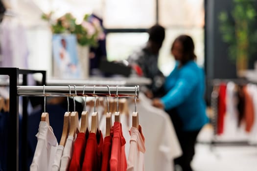 Selective focus of hangers full with trendy clothes in modern boutique, in background african american couple shopping for fashionable shirt, checking items fabric. Fashion concept
