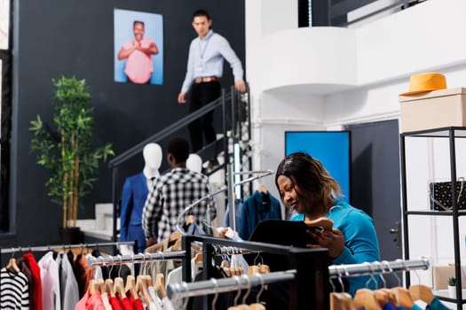 African american woman shopping for trendy outfit in clothing store, checking merchandise fabric. Shopaholic stylish customer wanting to buy new fashion collection in modern boutique