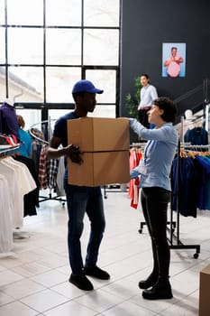 Store worker explaining customer shipping detalis to african american courier in modern boutique. Man wearing delivery uniform, holding cardboard boxes with online orders in shopping centre