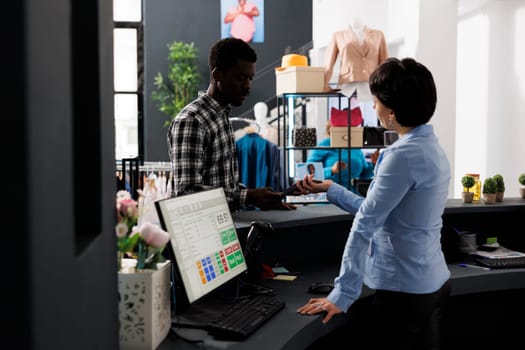 Stylish customer paying for fashionable clothes with credit card using payment terminal during fashion shopping session in modern boutique. African american man buying formal wear