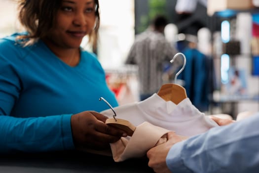 African american woman shopping for casual wear, holding multiple shirts in modern boutique. Stylish customer standing at counter desk, discussing items fabric with store employee. Fashion concept