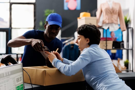 African american courier showing shipping report to worker, discussing online order in clothing store. Boutique manager standing at counter desk, preparing packages for delivery. Fashion concept