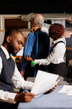 Teamworking tailors helping senior client find perfectly fitting bespoke business suit. Cheerful couturiers taking elderly gentleman measurements during sartorial customization comission process