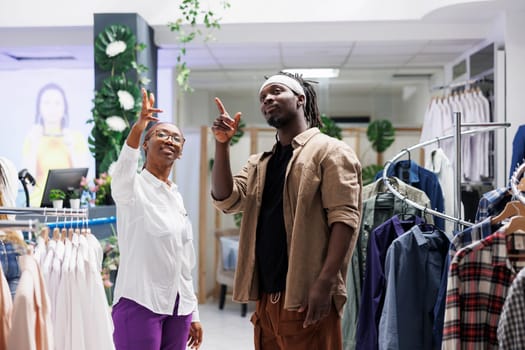 African american customer and store assistant pointing upwards while choosing trendy clothes. Man and woman examining stylish apparel purchase options in shopping mall boutique