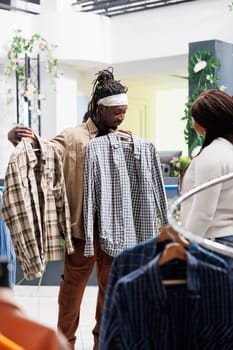 African american man showing woman two shirts to help with choice in clothing store. Customer holding apaprel on hangers and asking girlfriend for advice in fashion boutique