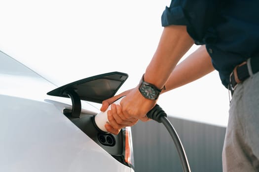 Young man put EV charger to recharge electric car's battery from charging station in city commercial parking lot. Rechargeable EV car for sustainable environmental friendly urban travel. Expedient