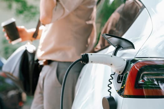 Young man with coffee cup, recharge electric car's battery from EV charging station in city commercial parking lot. Sustainable and eco friendly EV car with urban and shopping lifestyle. Expedient