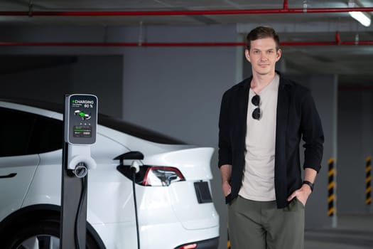 Young man travel with EV electric car to shopping center parking lot charging in downtown city showing urban sustainability lifestyle by green clean rechargeable energy of electric vehicle innards