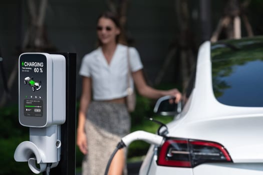 Young woman travel with EV electric car charging in green sustainable city outdoor garden in summer. Urban sustainability lifestyle by green clean rechargeable energy of electric BEV vehicle innards