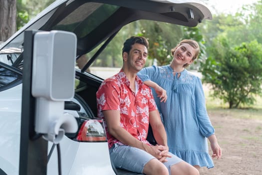 Road trip vacation with electric vehicle, lovely couple sitting on trunk and recharge EV car with green and clean energy. Nature and travel with eco-friendly car for sustainable environment. Perpetual