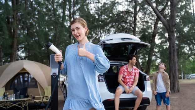 Outdoor adventure and family vacation camping at sea travel by eco friendly car. Cheerful woman or mother holding, pointing EV charger point with playful and happiness posture in campsite. Perpetual