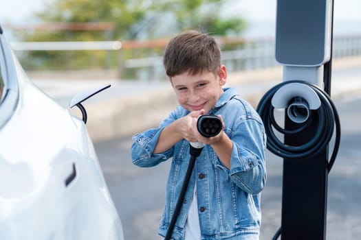 Playful little boy pointing EV charger at camera, recharging eco-friendly electric car from EV charging station. EV car travel by the seashore using clean and sustainable energy.Perpetual