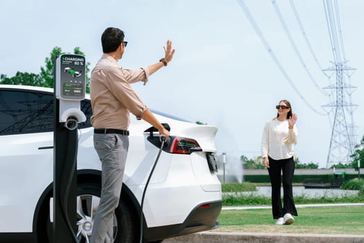 Young couple recharge EV car battery at charging station connected to power grid tower electrical industrial facility as electrical industry for eco friendly vehicle utilization. Expedient