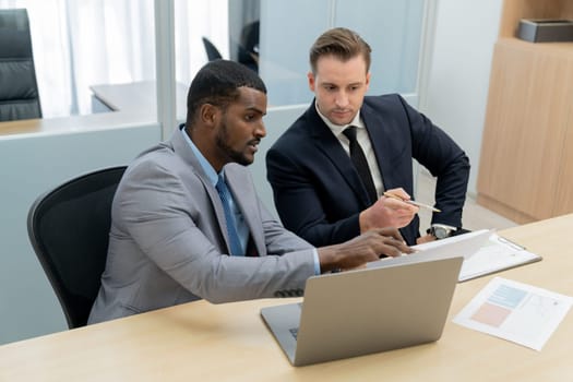 African male leader explain strategy while show solution to professional project manager by pointing at document, using laptop. Skilled business team brainstorming idea. Business meeting. Ornamented.