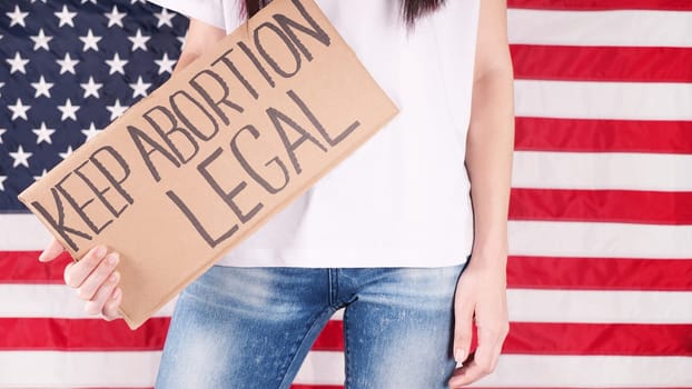 Young woman protester holds cardboard with Keep Abortion Legal sign against USA flag on background. Girl protesting against anti-abortion laws. Feminist power. Equal opportunity Womens rights reedom
