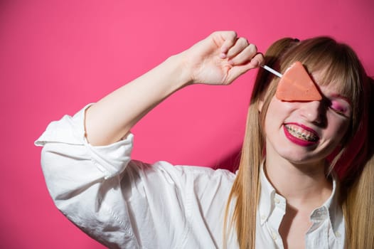 Portrait of a young woman with braces and bright makeup eating a lollipop on a pink background