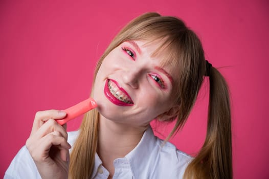 Portrait of a young woman with braces and bright makeup chewing gum on a pink background