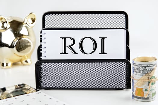 Return on investment concept. ROI text on a white piece of paper next to a piggy bank, a roll of money and a calculator on a white background