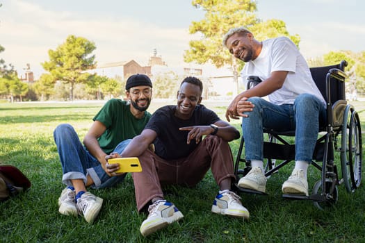 Young Black man in a wheelchair and his male friends taking selfie with phone sitting on the grass together in a park. Friendship and disability.