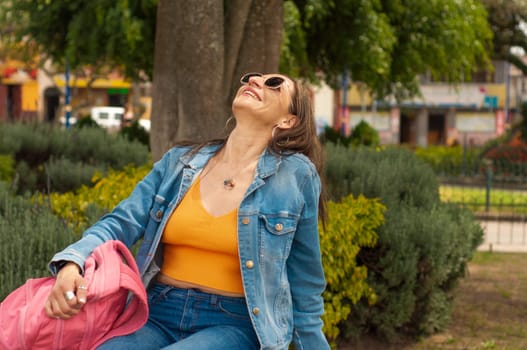 A woman, wearing jeans and a denim jacket, sits under a tree in the park, appearing cool and relaxed.