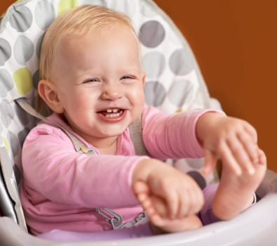 Happy, baby and laughing in high chair at home in the morning ready for eating food. Youth, funny and smile of a infant with development and laughter in a house with feet playing and joy of a child.