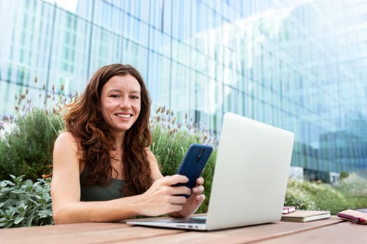 Female university student holding phone sitting on a table on college campus outdoors looking at camera. Copy space. Higher education and technology concept.