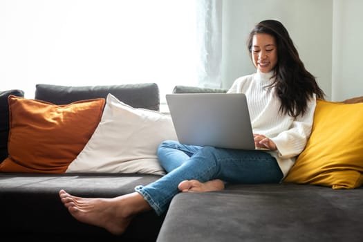 Happy smiling young Asian woman working with laptop at home sitting on the couch in cozy apartment living room. Copy space. Lifestyle concept.