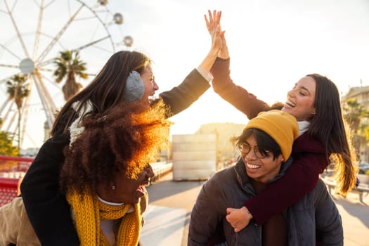 Happy multiracial young friends enjoying winter day outdoors. Boys giving piggyback to girls. Girls high five each other. Lifestyle concept.