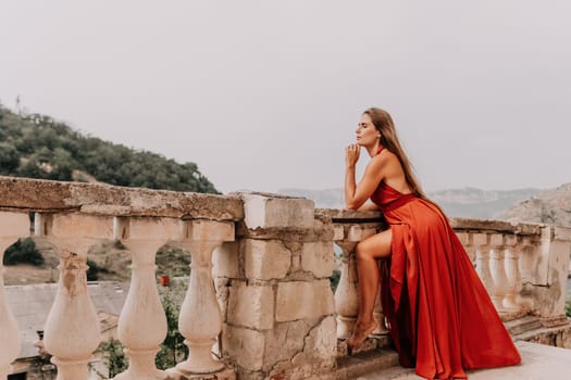 Side view a Young beautiful sensual woman in a red long dress posing on a volcanic rock high above the sea during sunset. Girl on the nature on overcast sky background. Fashion photo