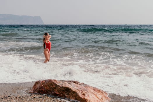 Woman travel sea. Young Happy woman in a long red dress posing on a beach near the sea on background of volcanic rocks, like in Iceland, sharing travel adventure journey