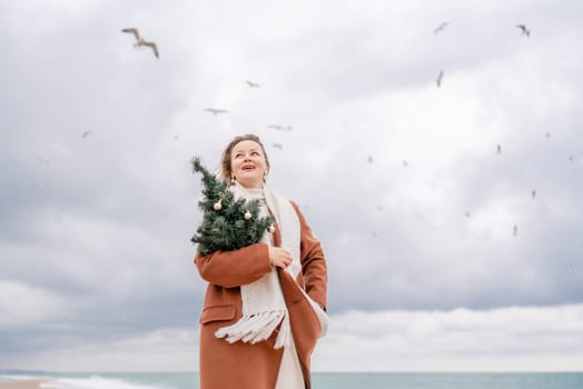 Blond woman Christmas sea. Christmas portrait of a happy woman walking along the beach and holding a Christmas tree on her shoulder. She is wearing a brown coat and a white suit