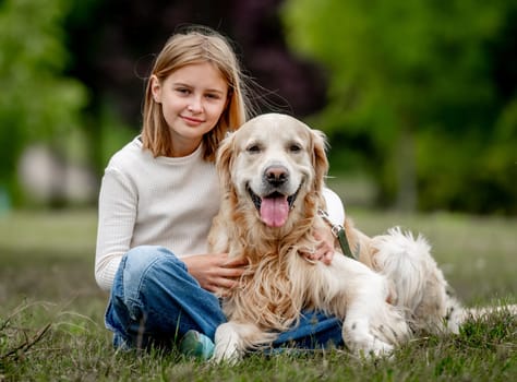 Preteen girl with golden retriever dog sitting at nature and looking back. Cute child kid hugging purebred pet doggy in park at summer