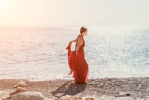 Side view a Young beautiful sensual woman in a red long dress posing on a rock high above the sea during sunrise. Girl on the nature on blue sky background. Fashion photo.