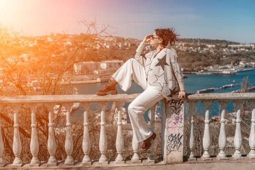 Woman walks around the city, lifestyle. A young beautiful woman in white trousers and a sweater sits on a white fence with balusters and overlooks the sea bay and the city