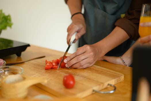 Young man cutting tomato on wooden board at kitchen counter. Closeup view.